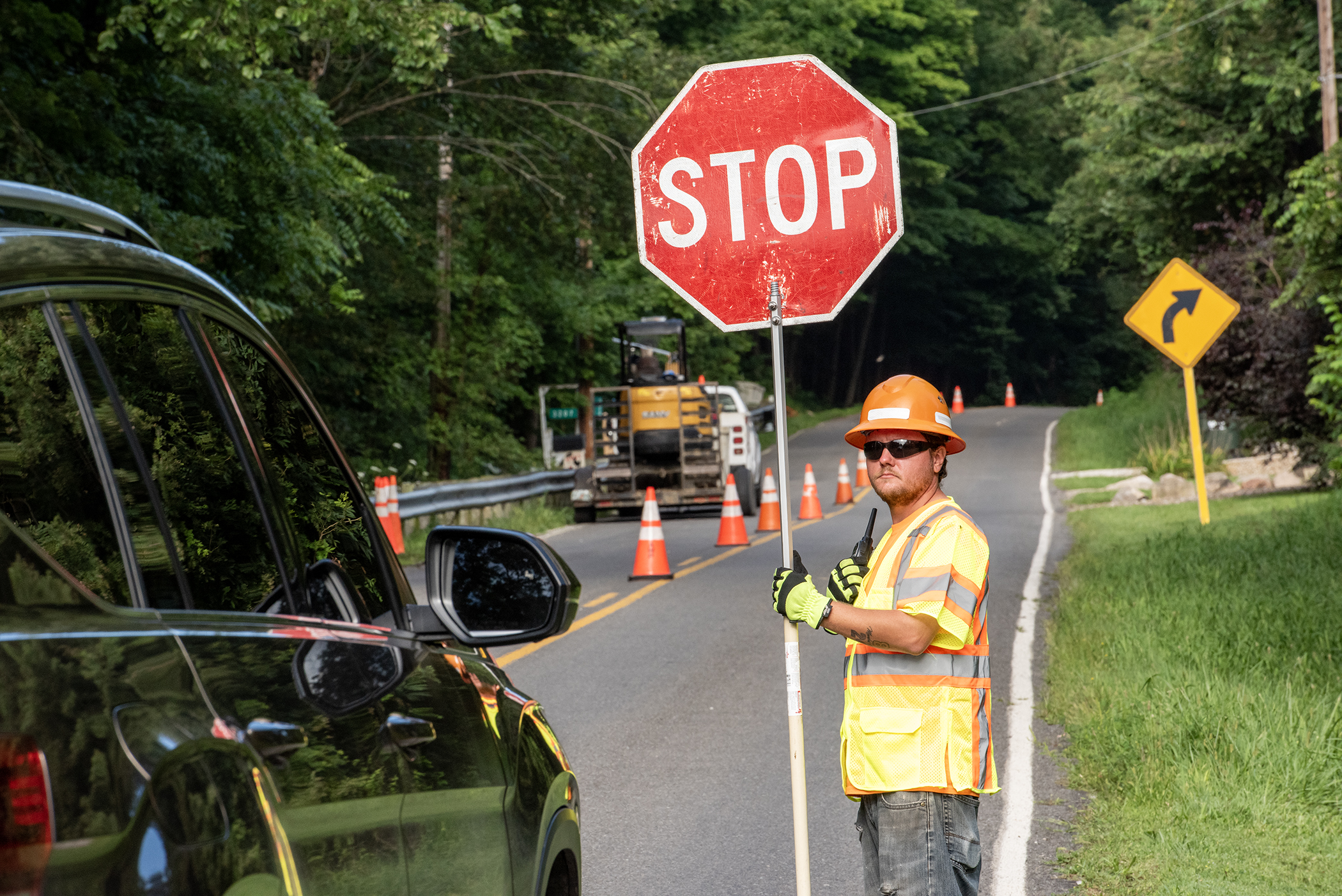 Construction worker in orange hi-vis vest holds a Stop sign on a road with cones and a yellow curve sign in the background.