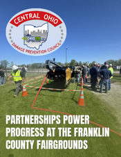People gathered at the Franklin County Fairgrounds around a vehicle with its trunk open, safety cones nearby, under a banner and the Central Ohio Damage Prevention Council seal.