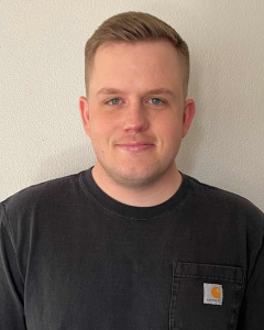 BRIDGER OBERG | OHIO811 Portrait of a man with short blond hair wearing a black T-shirt against a light wall, facing the camera with a slight smile.