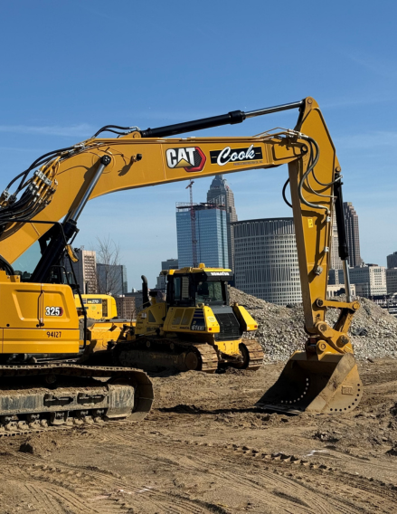 Yellow Caterpillar excavator with long arm and bucket at a construction site, urban skyline visible in the background.
