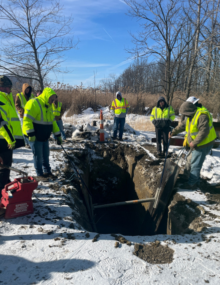 Snowy construction scene: several workers in high-visibility jackets stand around a deep open trench, inspecting the excavation with shovels and equipment nearby.