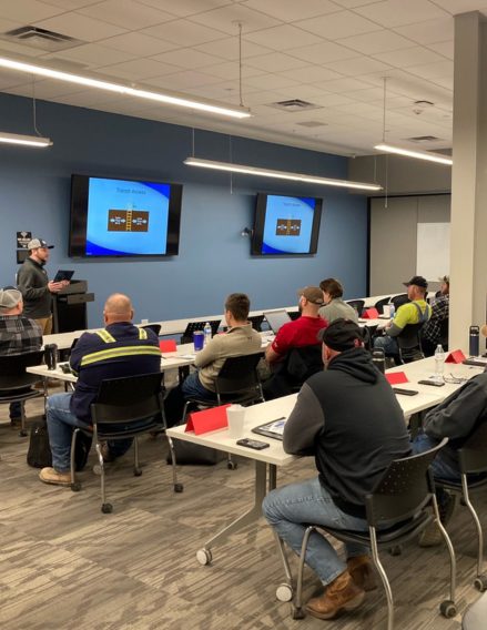 Instructor presenting at a training room front, two screens display slides as attendees sit at long tables with laptops and notebooks.