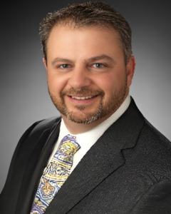 Chad Fritsch | OHIO811 Professional headshot of a smiling man in a dark suit and paisley-patterned tie, facing the camera with a neutral backdrop.