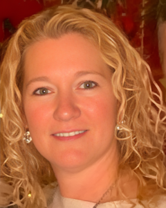 DEANNA DEWITT | OHIO811 Portrait of a smiling blonde woman with curly hair and earrings, close-up.