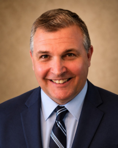 DERON LARGE | OHIO811 Professional headshot of a smiling man in a navy suit and striped tie against a neutral backdrop.
