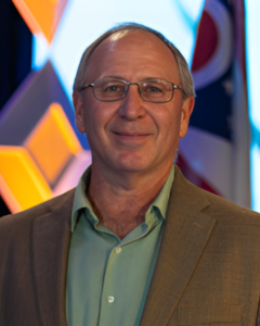 DON HUCK | OHIO811 Smiling middle-aged man with glasses in a green shirt and tan blazer, at an event with a colorful geometric backdrop.