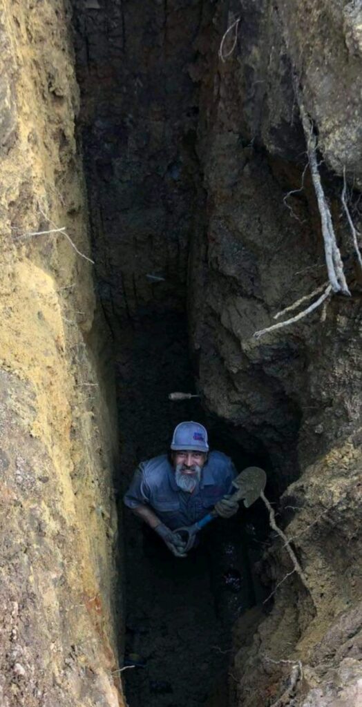 | OHIO811 Man with gray beard in a blue work shirt and cap standing in a deep, narrow trench, holding a shovel.
