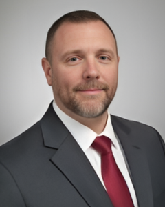 JUSTIN FREUDEMAN | OHIO811 Professional headshot of a man in a dark suit and red tie, neutral gray background, smiling slightly at the camera.