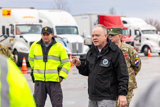 Man in a black jacket speaks to uniformed staff in a truck yard briefing session