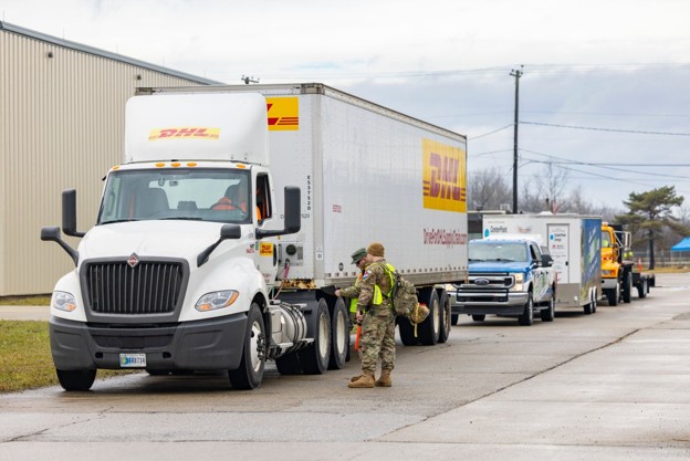 Soldier in camouflage and a safety vest directs a DHL tractor-trailer at a loading area with a line of DHL trucks nearby.