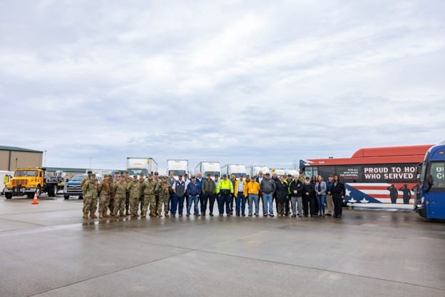 Group of soldiers and civilians posing on a wet tarmac with buses and trucks behind them on an airfield.