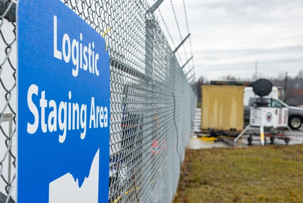 Blue sign reading 'Logistic Staging Area' on a chain-link fence at an industrial site, with equipment in the background.