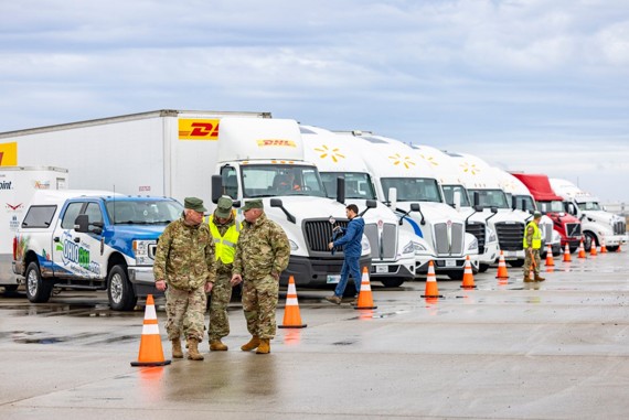 Military personnel in camouflage discuss next to a line of delivery trucks and vans behind orange traffic cones in a large parking lot during a convoy drill or inspection moment.