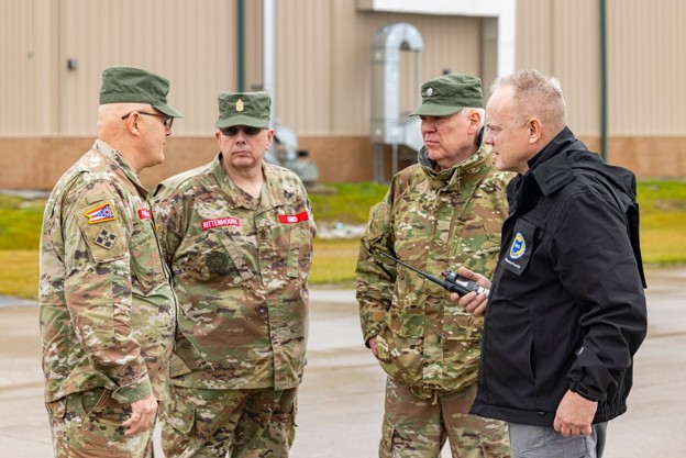 Four soldiers in camouflage uniforms stand outside a beige hangar, conversing with a civilian official who holds a radio.