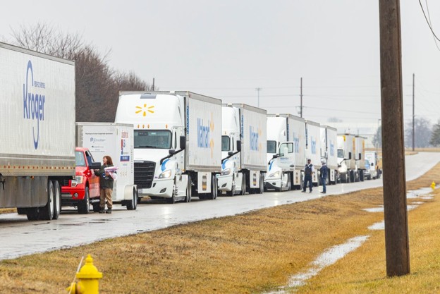 Line of Kroger delivery trucks lined up along a wet road, with a red car and workers standing near the first trucks.