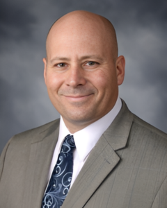 MICHAEL WILSON | OHIO811 Professional headshot: bald man in a gray suit and blue patterned tie, smiling against a neutral studio background.