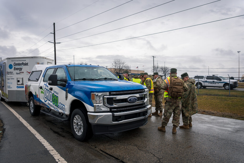 Group of military personnel and utility workers in high-visibility vests standing near a blue CenterPoint Energy service truck in a parking lot, with a police SUV in the background.
