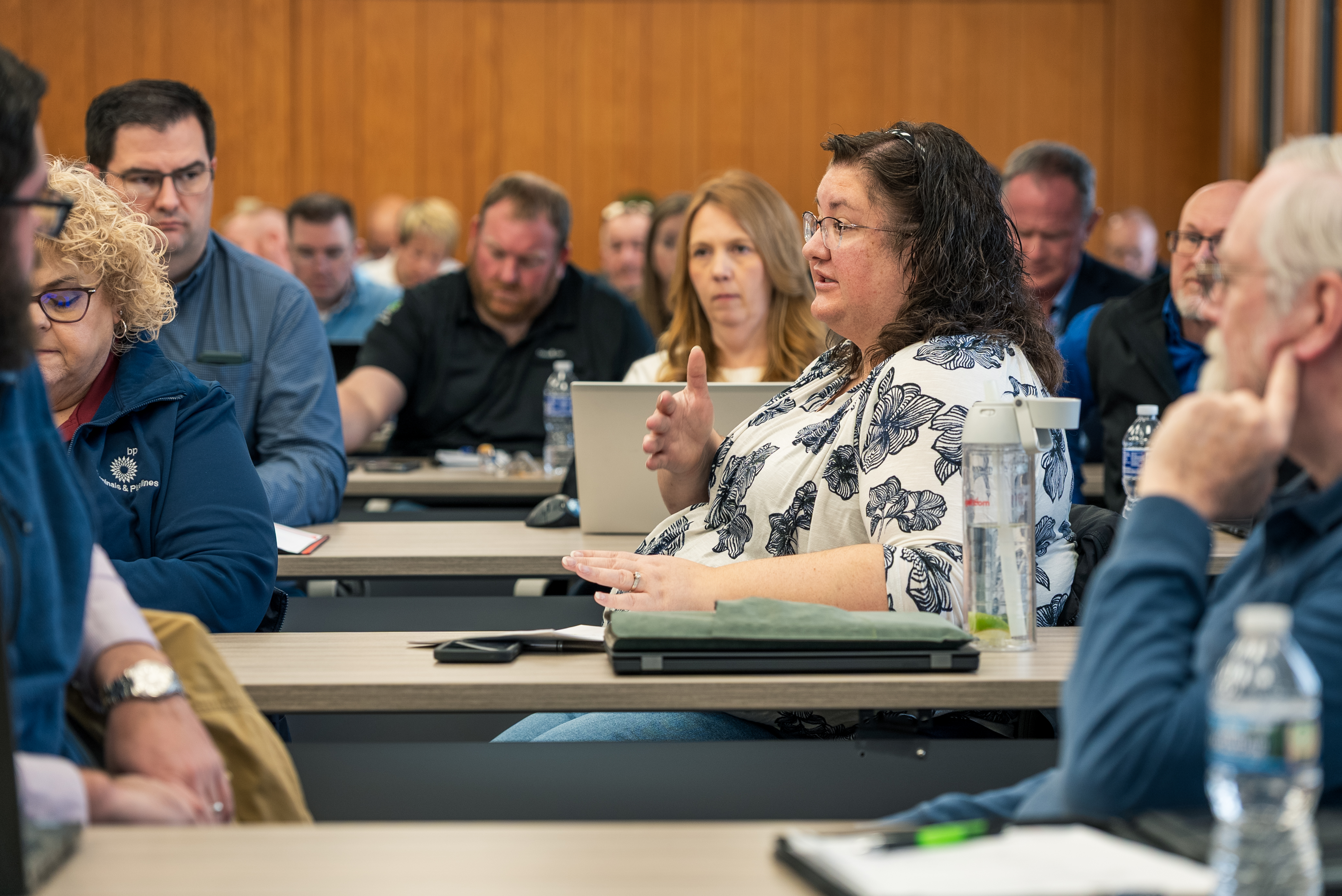 Woman with dark hair and glasses speaks during a meeting, others listening at a long table in a conference room.