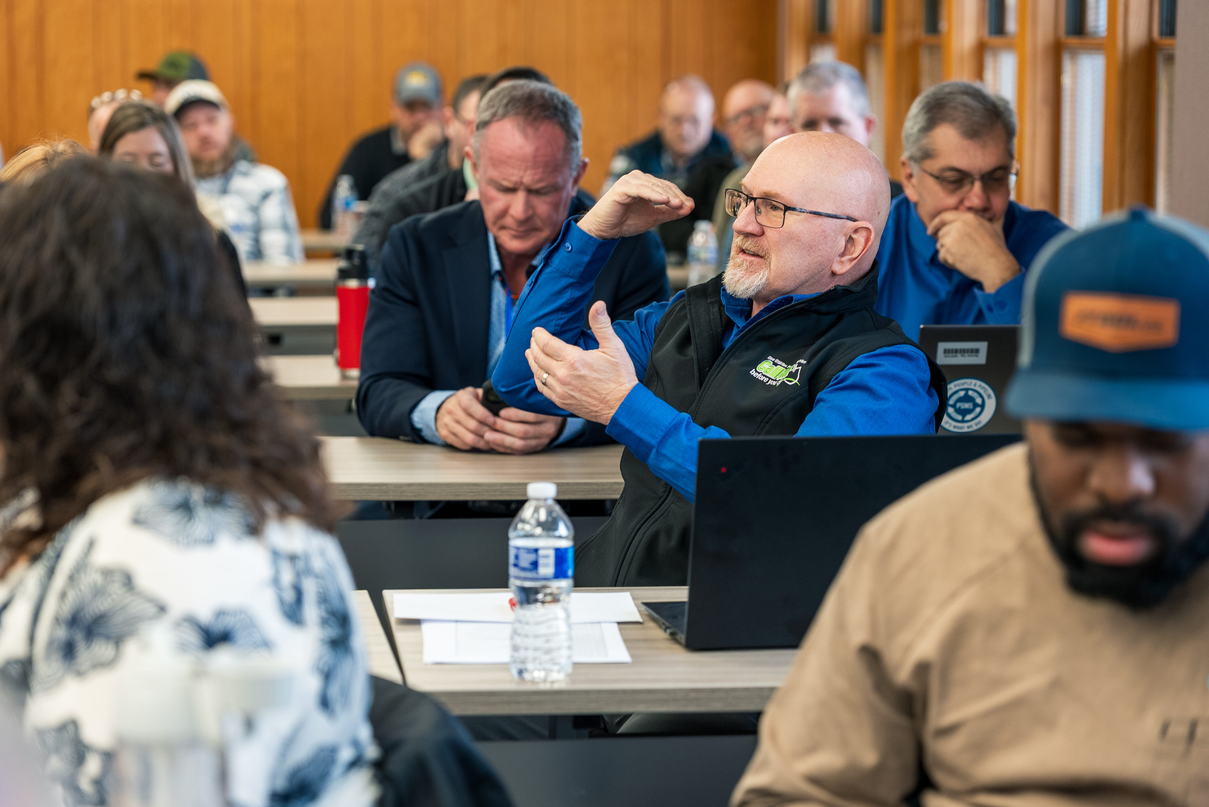 Man in blue shirt gestures with hands while speaking at a classroom table, others listening in background; water bottle and laptops visible.