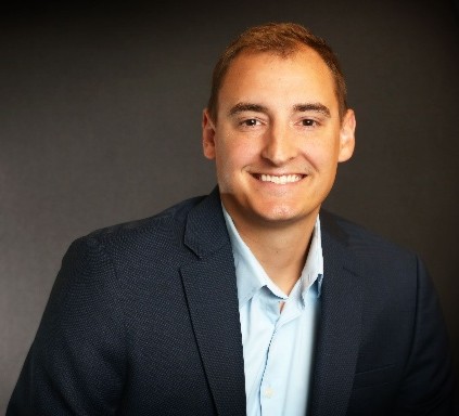 Head-and-shoulders portrait of a smiling man in a navy blazer and light blue shirt against a dark backdrop.