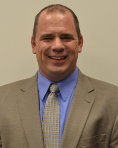 THOMAS TRAVIS | OHIO811 Professional headshot of a smiling man wearing a tan suit, light blue shirt, and patterned tie.