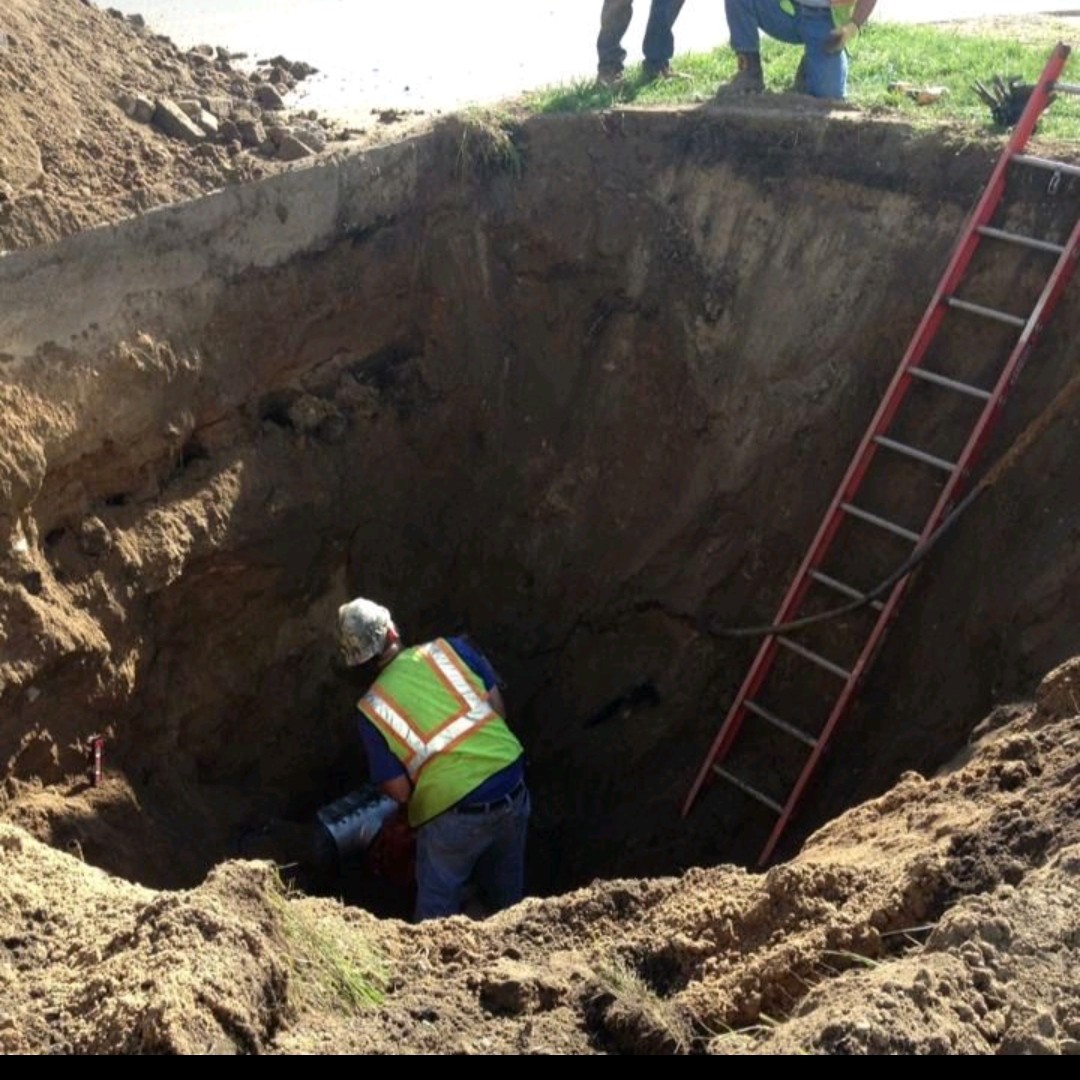 | OHIO811 Worker in a high‑visibility vest and hard hat kneels inside a deep dirt trench, with a red ladder leaning along the wall nearby on a construction site.