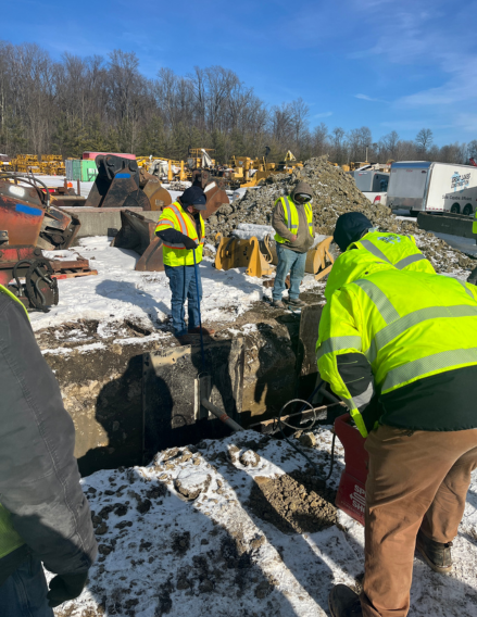 Construction crew in neon vests standing around a deep trench with dirt and concrete, snow on the ground and heavy equipment nearby.