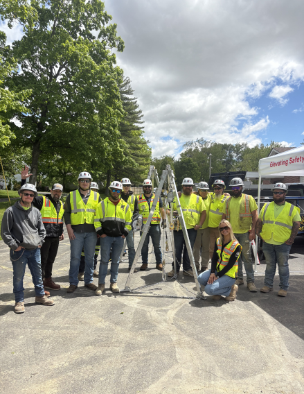 Construction crew in safety vests and hard hats posing around a survey tripod on a paved site outdoors.