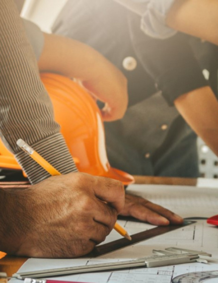 Close-up of a worker's hands drawing on a blueprint with a pencil, orange hard hat nearby on the table.