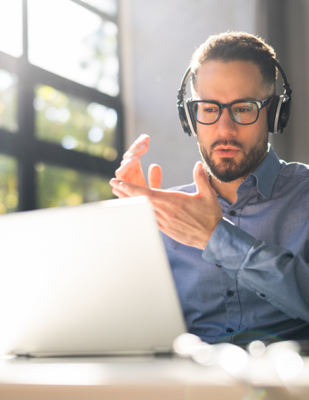 Bearded man wearing headphones and glasses sits at a laptop, gesturing with his hands as he talks.
