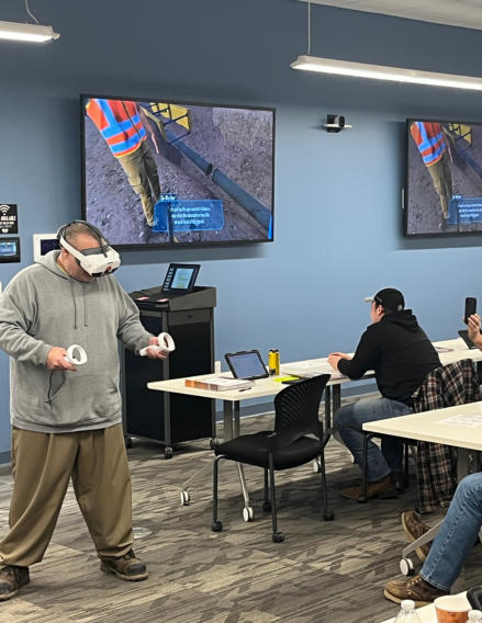 Man wearing a virtual reality headset and holding motion controllers stands in a training room, while two people at desks use laptops and safety training screens are visible on the wall.