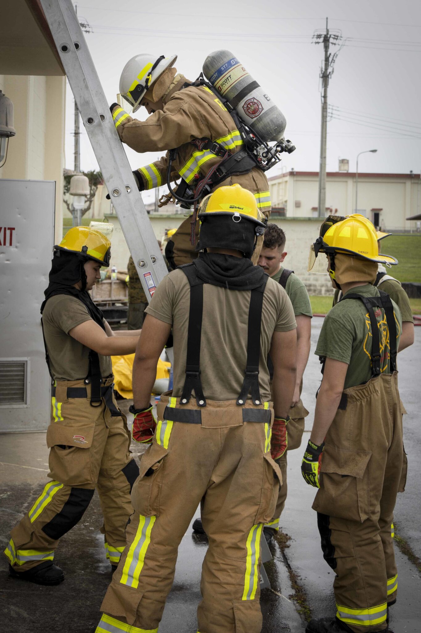 Military firefighters conduct the Physical Abilities Test | OHIO811 Firefighters in tan turnout gear with reflective stripes position a ladder during training outdoors on a wet pavement.