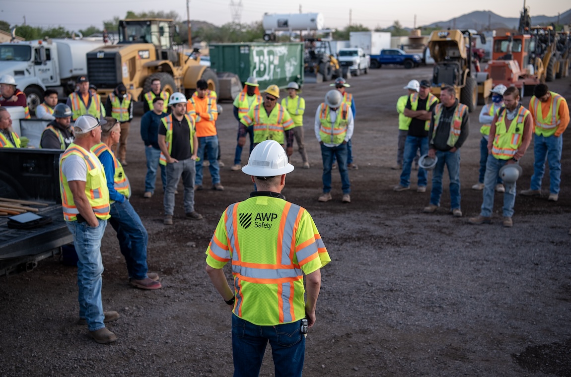 AWP_1 | OHIO811 Supervisor in a white helmet and neon safety vest addresses a circle of workers at a gravel construction site.