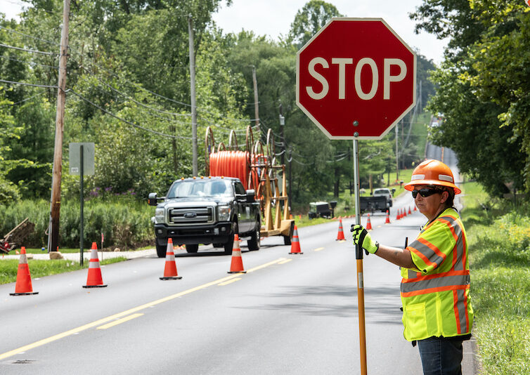 AWP_3 | OHIO811 Construction worker standing on a road holding a stop sign with orange cones and construction equipment ahead.