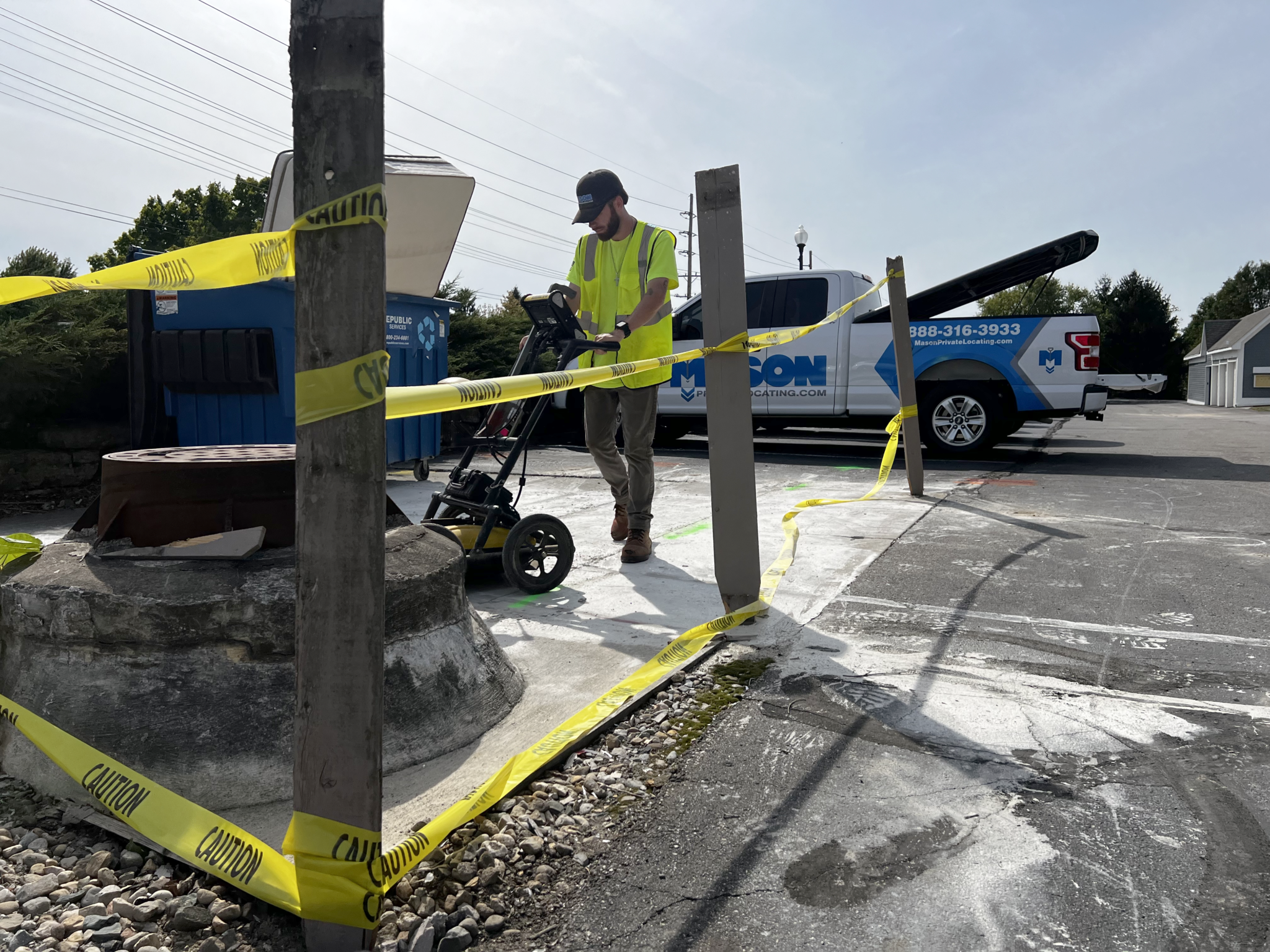 Mason howard job | OHIO811 Construction worker in a neon shirt operates equipment behind yellow caution tape near a blue dumpster and a pickup truck with its bed raised in the background.